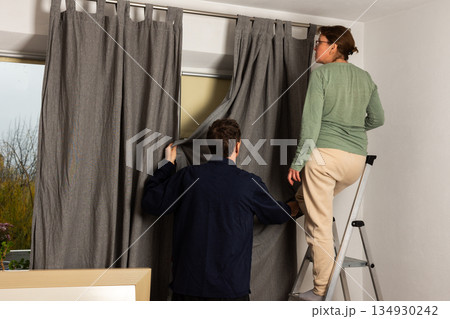 A married couple works together to hang freshly washed curtains in their living room. They position the curtains as they prepare for the autumn season in their home 134930242