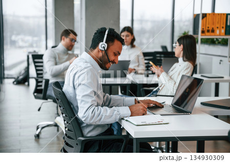 Man is sitting by laptop in headphones. Four people are working in the office together 134930309