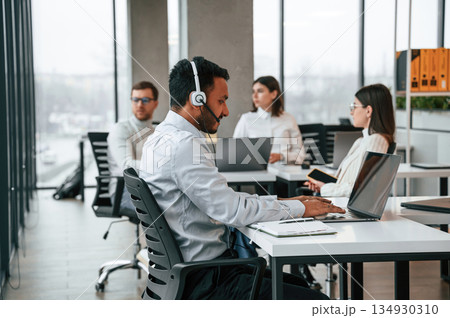 Man is sitting by laptop in headphones. Four people are working in the office together Man is sitting by laptop in headphones. Four people are working in the office together 134930310