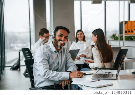 Man in headphones is sitting and smiling. Four people are working in the office together Man in headphones is sitting and smiling. Four people are working in the office together 134930311