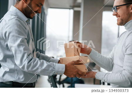 View from the side. Holding paper eco boxes. Two men are working in the modern office together View from the side. Holding paper eco boxes. Two men are working in the modern office together 134930373