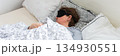 A 50-year-old woman is sound asleep in her bed, wearing a sleep mask. She is surrounded by pillows and blankets, creating a tranquil atmosphere. Sunlight gently filters into the room, banner 134930551