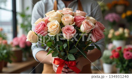 A florist presenting a bouquet of pink and cream roses tied with a red ribbon, in a shop filled with vibrant floral arrangements. Flower delivery concept 134931240