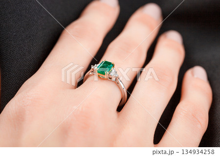 Beautiful female hand with gold ring with diamonds and emerald on gray background Beautiful female hand with gold ring with diamonds and emerald on gray background 134934258