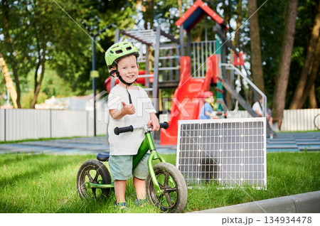 Young boy beside solar panel on the grass, gives thumbs-up. Smiling child with green balance bike. Behind him, playground with red slides and climbing structures surrounded by trees and fence. 134934478
