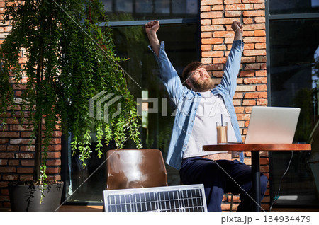 Man stretches with arms overhead while working on laptop at outdoor round table. Solar panel nearby, emphasizing sustainable, modern eco-friendly workspace that combines technology and sustainability. 134934479