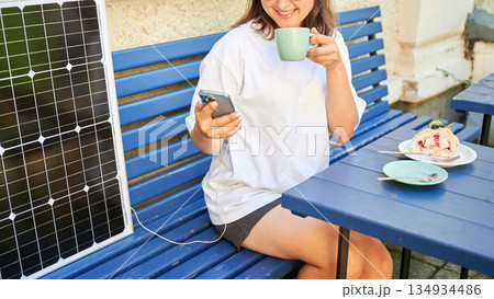 Girl drinking coffee while phone charging from solar panel. Cropped view of happy woman sitting next to portable solar battery charger. Integration of sustainable renewable energy into everyday life. 134934486