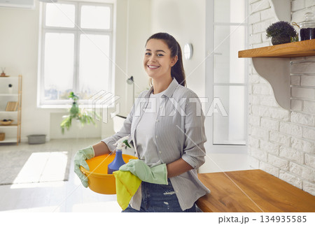 Woman housewife in gloves with cleaning tools and rags, standing in kitchen and looking at camera. Woman housewife in gloves with cleaning tools and rags, standing in kitchen and looking at camera. 134935585