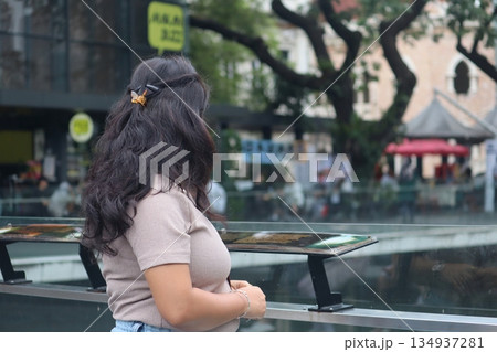 Woman observing a city plaza fountain in Kuala Lumpur, Malaysia, surrounded by trees, cafes, and everyday urban activity. Woman observing a city plaza fountain in Kuala Lumpur, Malaysia, surrounded by trees, cafes, and everyday urban activity. 134937281