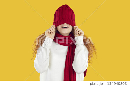 Funny teenage girl in scarf behaves cheerfully and playfully, pulling red warm hat on her eyes. Funny teenage girl in scarf behaves cheerfully and playfully, pulling red warm hat on her eyes. 134940808