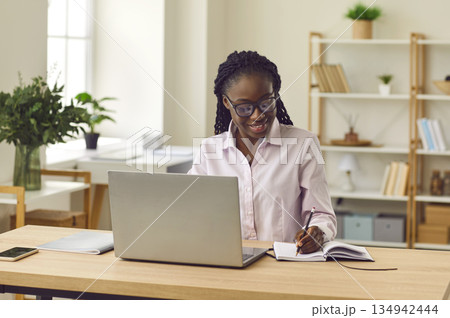 Happy African American student girl sitting at table, using laptop, and taking notes Happy African American student girl sitting at table, using laptop, and taking notes 134942444
