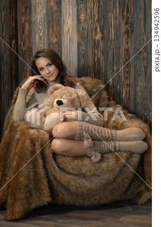 Young woman sits on a chair holding a teddy bear in a cozy indoor space with wood walls Young woman sits on a chair holding a teddy bear in a cozy indoor space with wood walls 134942596