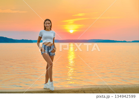 Young woman enjoying a sunset by the lake while wearing a white top and denim shorts Young woman enjoying a sunset by the lake while wearing a white top and denim shorts 134942599