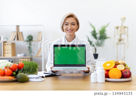 A smiling nutritionist holds a laptop with a green screen, surrounded by fruits and vegetables, promoting healthy eating. A smiling nutritionist holds a laptop with a green screen, surrounded by fruits and vegetables, promoting healthy eating. 134943135