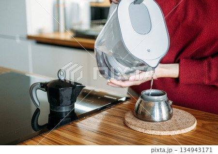 Person pours filtered water from pitcher into base of moka pot on wooden counter. Manual espresso routine, elevated home brewing, artisanal coffee gear, precision coffee making. 134943171