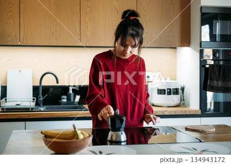 Woman operates induction stove to brew coffee with moka pot in modern kitchen. Induction stove brewing, modern kitchen appliances, smart home living. 134943172