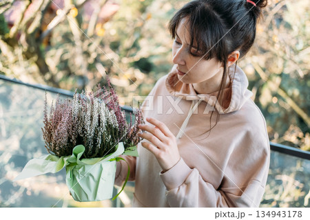Woman examines potted heather plant on sunlit balcony. Biophilia, indoor outdoor living, balcony plants, heather flower. Woman examines potted heather plant on sunlit balcony. Biophilia, indoor outdoor living, balcony plants, heather flower. 134943178