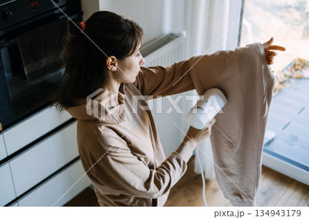 Woman uses handheld garment steamer on trousers in kitchen. Smart home appliances, time-saving technology, modern laundry care. 134943179