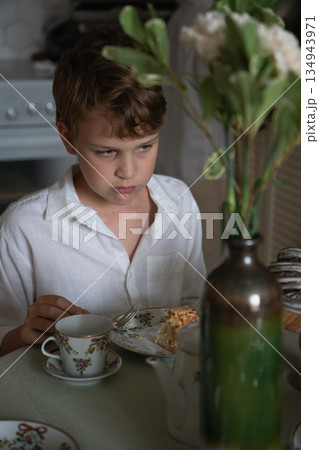 Young boy sits at home table with a cup of tea and a slice of cake, showing a thoughtful mood in soft natural light and a cozy kitchen setting 134943971