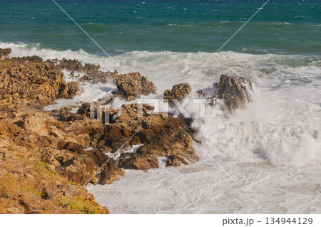 Sea foam crashing against rocky shore. 134944129