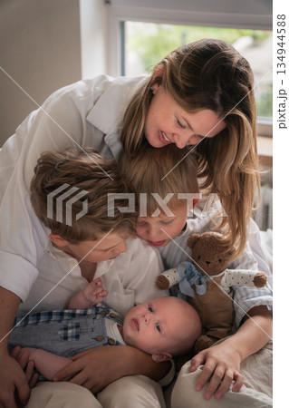 A mother embraces her three young sons, smiling warmly by the window as they sit together on the bed 134944588