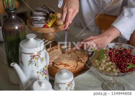Hands cut a homemade apple charlotte on a tea table with floral teapot, cups, cookies, and bowls of red and white currants in soft natural light Hands cut a homemade apple charlotte on a tea table with floral teapot, cups, cookies, and bowls of red and white currants in soft natural light 134944608