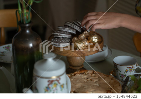 Hand reaches for sweets on a wooden stand beside homemade apple charlotte, gingerbread cookies, candies, and a floral tea set on a cozy table 134944622
