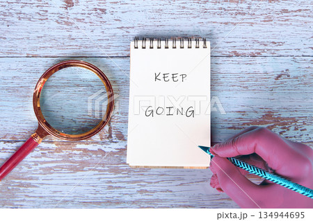 On a rustic wooden surface, a hand holds a pencil next to a notepad displaying the phrase KEEP GOING, accompanied by a magnifying glass providing encouragement and focus On a rustic wooden surface, a hand holds a pencil next to a notepad displaying the phrase KEEP GOING, accompanied by a magnifying glass providing encouragement and focus 134944695