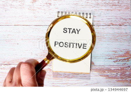 A hand holds a magnifying glass over a notepad. The notepad displays the text STAY POSITIVE. This rests on a faded wooden surface, creating a scene of encouragement 134944697