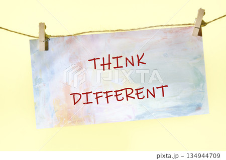 A colorful paper with the words Think Different written in red ink hangs by clothespins on a string. The paper has a painted watercolor effect 134944709