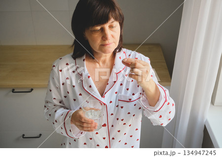 Mature Woman taking medication with glass of water in her pajamas at home Mature Woman taking medication with glass of water in her pajamas at home 134947245