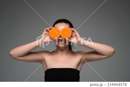 Woman holding fresh orange halves over eyes on neutral background, creative healthy lifestyle portrait with citrus fruit, vitamins, wellness and natural beauty 134948578