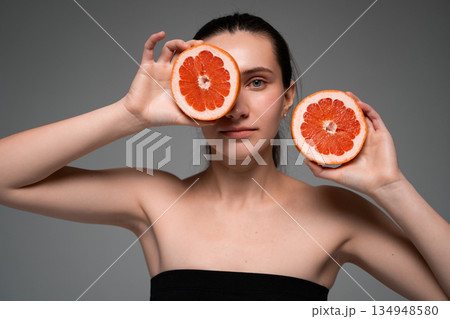 Smiling woman holding fresh grapefruit halves near face on neutral background, healthy lifestyle, natural beauty, vitamins, wellness concept 134948580