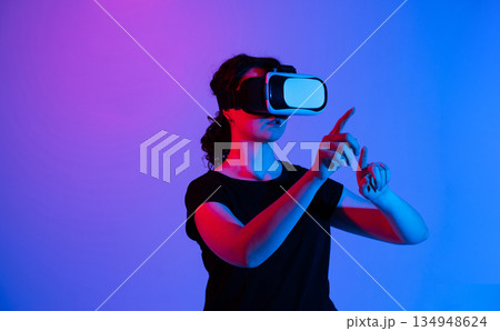 Woman in virtual reality headset interacting with invisible digital interface using hand gestures with copy space and neon background Woman in virtual reality headset interacting with invisible digital interface using hand gestures with copy space and neon background 134948624
