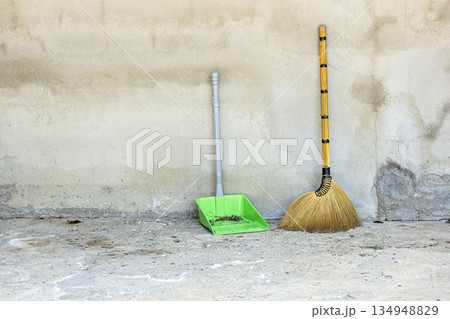 A traditional grass broom with a golden handle and a modern green plastic dustpan, holding debris, lean against a textured A traditional grass broom with a golden handle and a modern green plastic dustpan, holding debris, lean against a textured 134948829