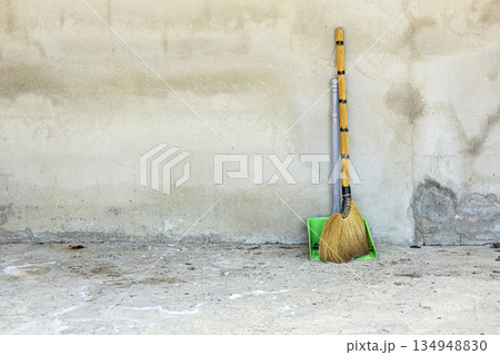 A traditional grass broom with a black-wrapped handle and a bright green plastic dustpan lean against a textured A traditional grass broom with a black-wrapped handle and a bright green plastic dustpan lean against a textured 134948830