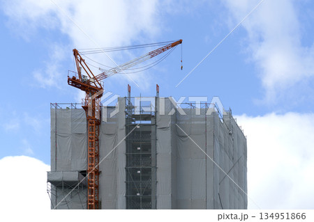Large orange tower crane on top of a building under construction wrapped in grey safety mesh. Skyscraper development project against a bright blue sky with white clouds. Urban growth concept. Large orange tower crane on top of a building under construction wrapped in grey safety mesh. Skyscraper development project against a bright blue sky with white clouds. Urban growth concept. 134951866