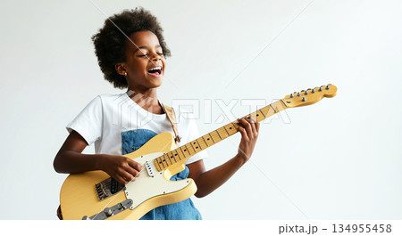 Young girl with curly hair joyfully playing electric guitar, wearing casual clothing, in a bright studio setting, showcasing musical talent and passion for music 134955458