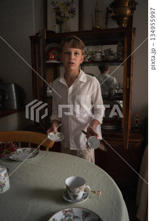 Young boy helping to set the table at home, holding a cup and saucer, with his reflection visible in a glass cabinet behind him 134955997