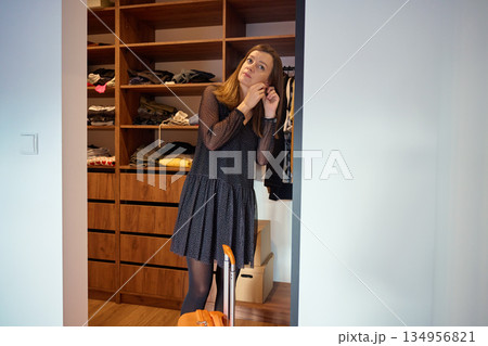 Woman standing near wardrobe wearing dress and putting on earrings with suitcase nearby 134956821