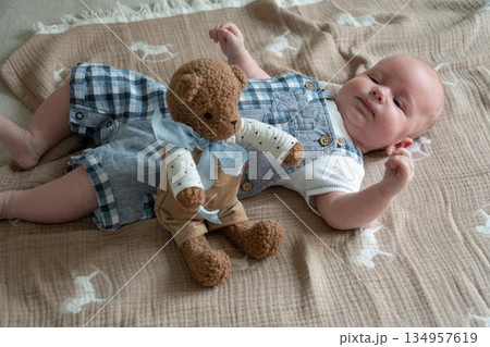 Newborn baby lies on a soft blanket with a plush teddy bear, neutral tones, gentle daylight, calm and cozy infant moment at home 134957619
