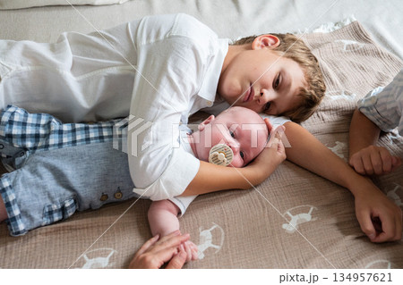 Two brothers lie on a bed hugging a newborn baby with a pacifier, soft daylight, neutral colors, calm and tender sibling bonding at home 134957621