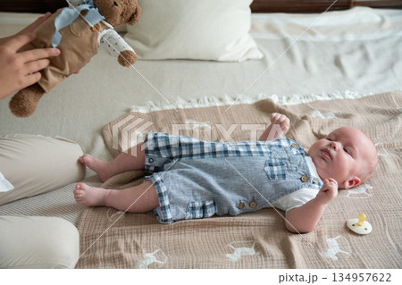 Newborn baby lies on soft blanket on bed while a hand holds a plush teddy bear nearby, neutral colors, gentle daylight, calm and cozy home atmosphere 134957622