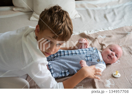 Older brother gently touches a newborn baby lying on a bed at home, soft daylight, neutral colors, tender sibling interaction 134957708