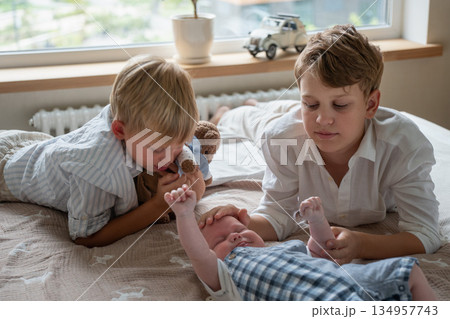 Two older brothers lying on bed, gently interacting with newborn baby in a bright home interior, showing care, tenderness, sibling bonding Two older brothers lying on bed, gently interacting with newborn baby in a bright home interior, showing care, tenderness, sibling bonding 134957743