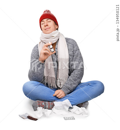 Sick man in winter hat holding nasal spray and feeling relief, sitting with tissues and pills isolated on white background. High quality photo 134958121