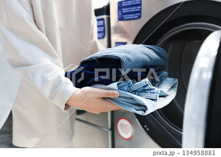 Woman holding stack of clean jeans near washing machine. Close up of hands with laundry at laundromat. High quality photo 134958881
