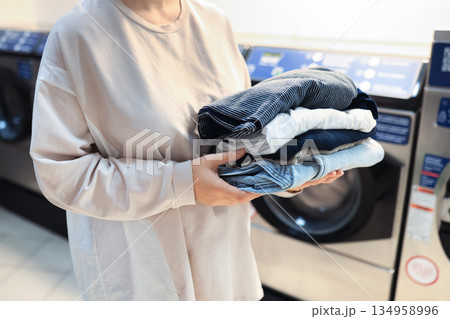 Woman holding stack of folded clean clothes at public laundromat with washing machines in background. High quality photo 134958996