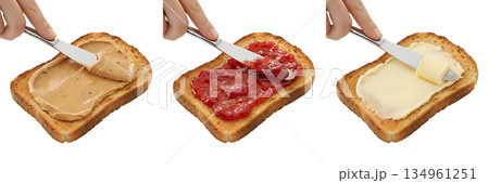Process of spreading peanut butter, strawberry jam, and butter on slices of toasted bread to make a classic sandwich, isolated 134961251