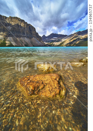 Bow Lake Crystal Water and Rocky Shore with Dramatic Mountain Range Under Cloudy Sky 134965737